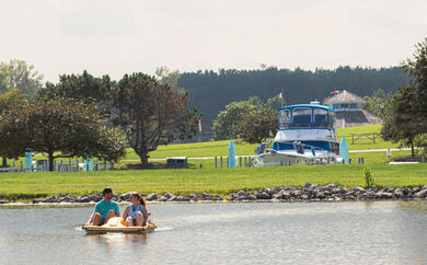 Two people in a paddleboat in the water, with a large boat on land in the background.